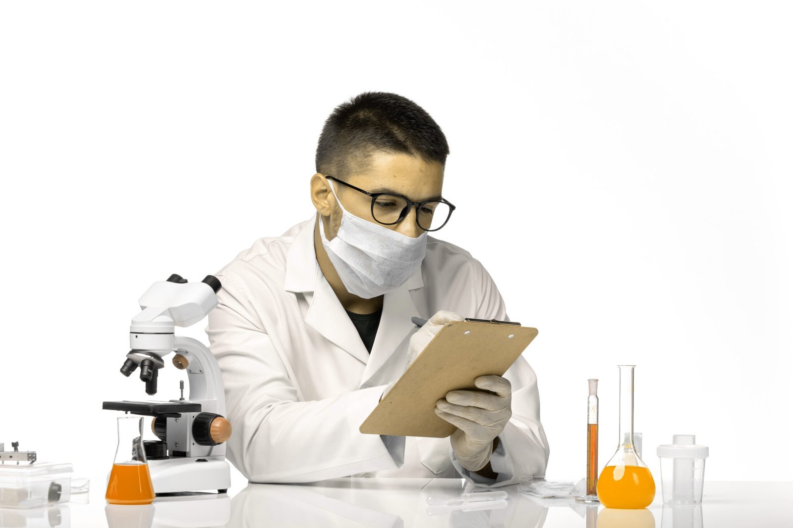 A researcher in a lab coat and mask writing notes beside a microscope
